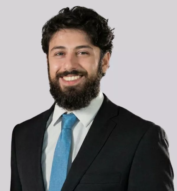 Smiling man with dark hair and beard in a black suit and blue tie, posing against a light gray background.