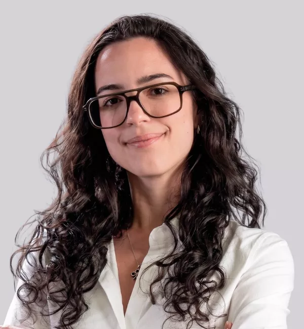 Woman with long dark curly hair and glasses, wearing a white blouse, smiling softly against a gray background.