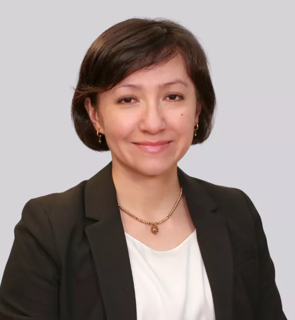 Portrait of a woman in a black blazer and white blouse, smiling softly, wearing a gold necklace and small earrings, against a light gray background.