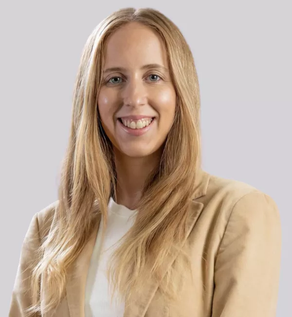 Portrait of a smiling woman with long blonde hair wearing a beige blazer and white top against a light gray background.