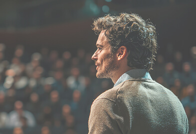 Male presenter on stage in profile, addressing a crowd in a dimly lit venue