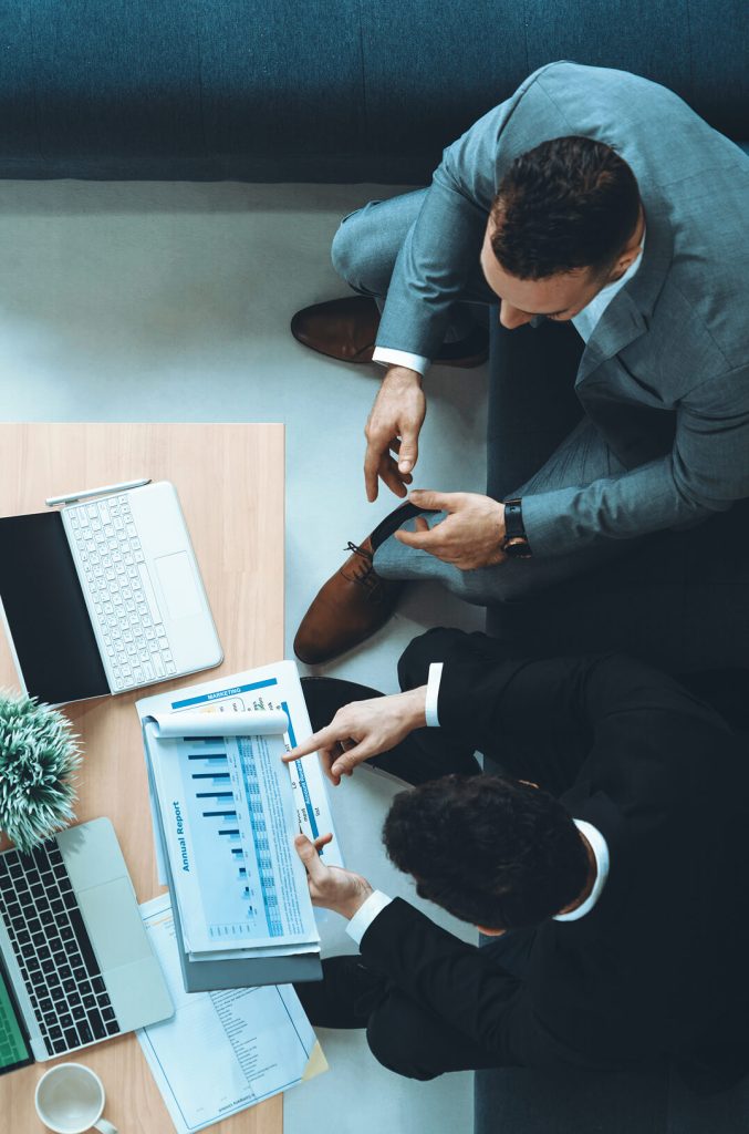 Top-down view of two professionals in suits reviewing a financial chart and annual report at a desk with a laptop nearby.